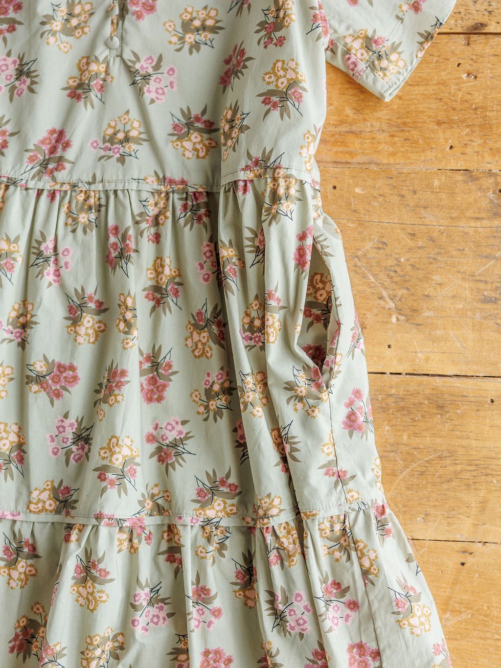 A close-up of the Tularosa Floral Dress by Savannah Sevens western life{&}style, featuring sage cotton with pink and yellow flowers, short sleeves, ruffle trim, gathered waist, and a side pocket, laid out on a wooden surface.