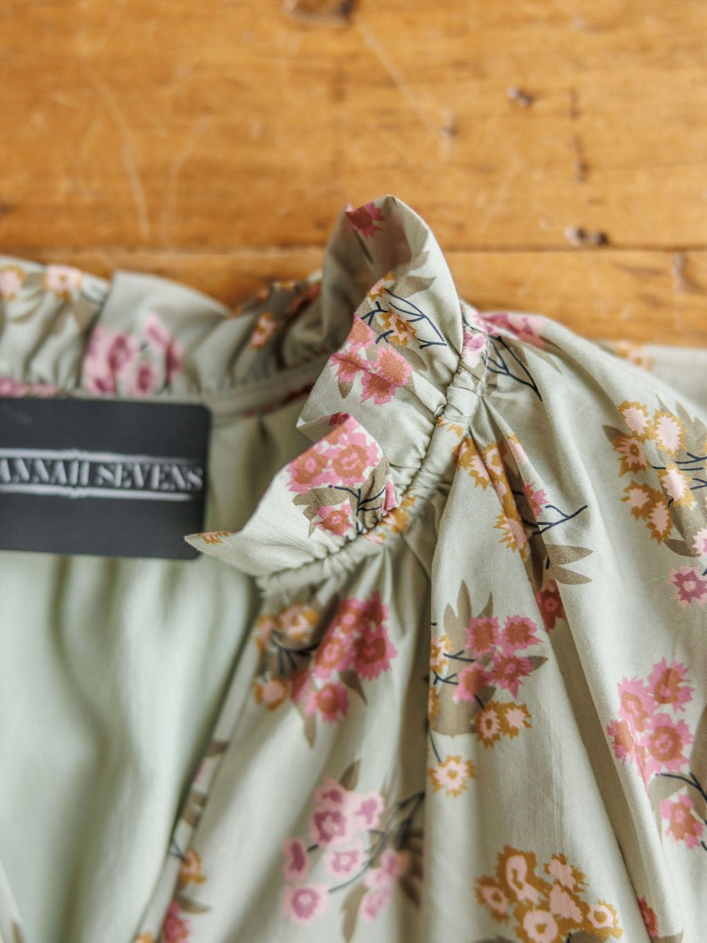 A close-up of the Tularosa Floral Dress by Savannah Sevens western life{&}style, featuring a sage base, pink and yellow floral print, ruffled neckline, and black tag, displayed on a wooden surface.