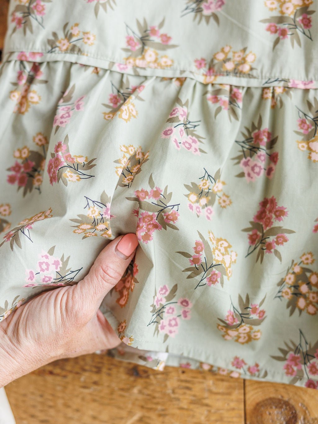 A hand with pink nail polish holds the hem of the Tularosa Floral Dress by Savannah Sevens western life{&}style, featuring a sage ruffle design with pink, yellow, and green floral print, spread out on a wooden surface.