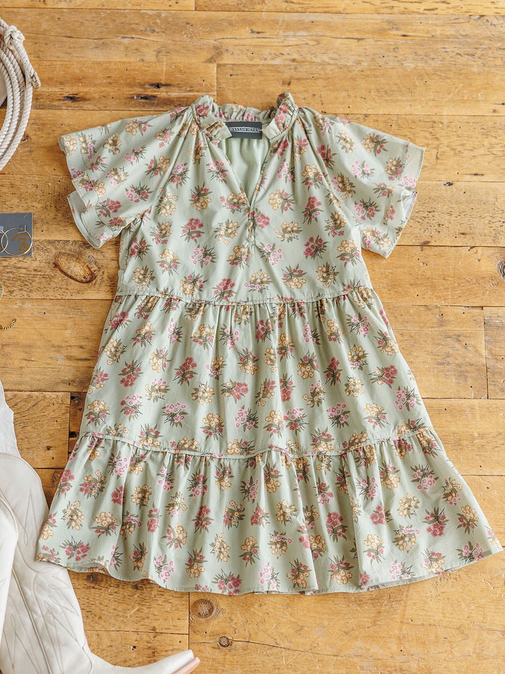 The Tularosa Floral Dress by Savannah Sevens western life{&}style—a light green cotton short sleeve dress with a tiered skirt and small yellow, pink, and green flowers—lies flat on a wooden floor. White boots peek in from the left.