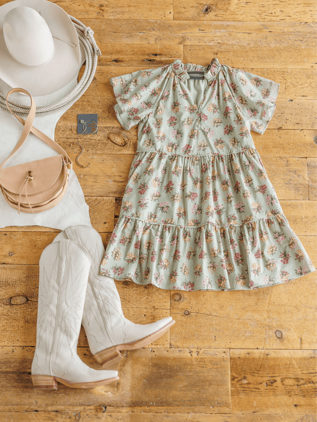 The Tularosa Floral Dress by Savannah Sevens western life{&}style is styled on a wooden floor with white cowboy boots, a beige hat, tan crossbody bag, and gold jewelry for a casual Western-inspired look.