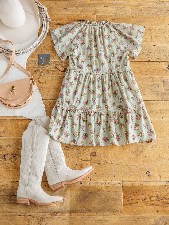 The Tularosa Floral Dress by Savannah Sevens western life{&}style is styled on a wooden floor with white cowboy boots, a beige hat, tan crossbody bag, and silver jewelry for a chic western-inspired look.