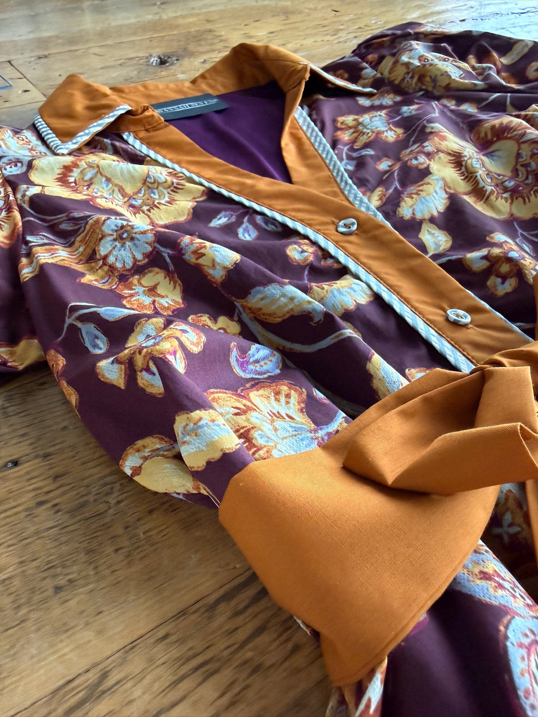 Presenting the Tullahassee Floral Dress from Savannah Sevens western life{&}style: a button-up dress crafted from rich purple fabric patterned with yellow and blue florals. It features a solid mustard yellow collar, placket, and wide cuffs for contrast, all accented by a blue-and-white pinstriped trim along the collar and front opening.