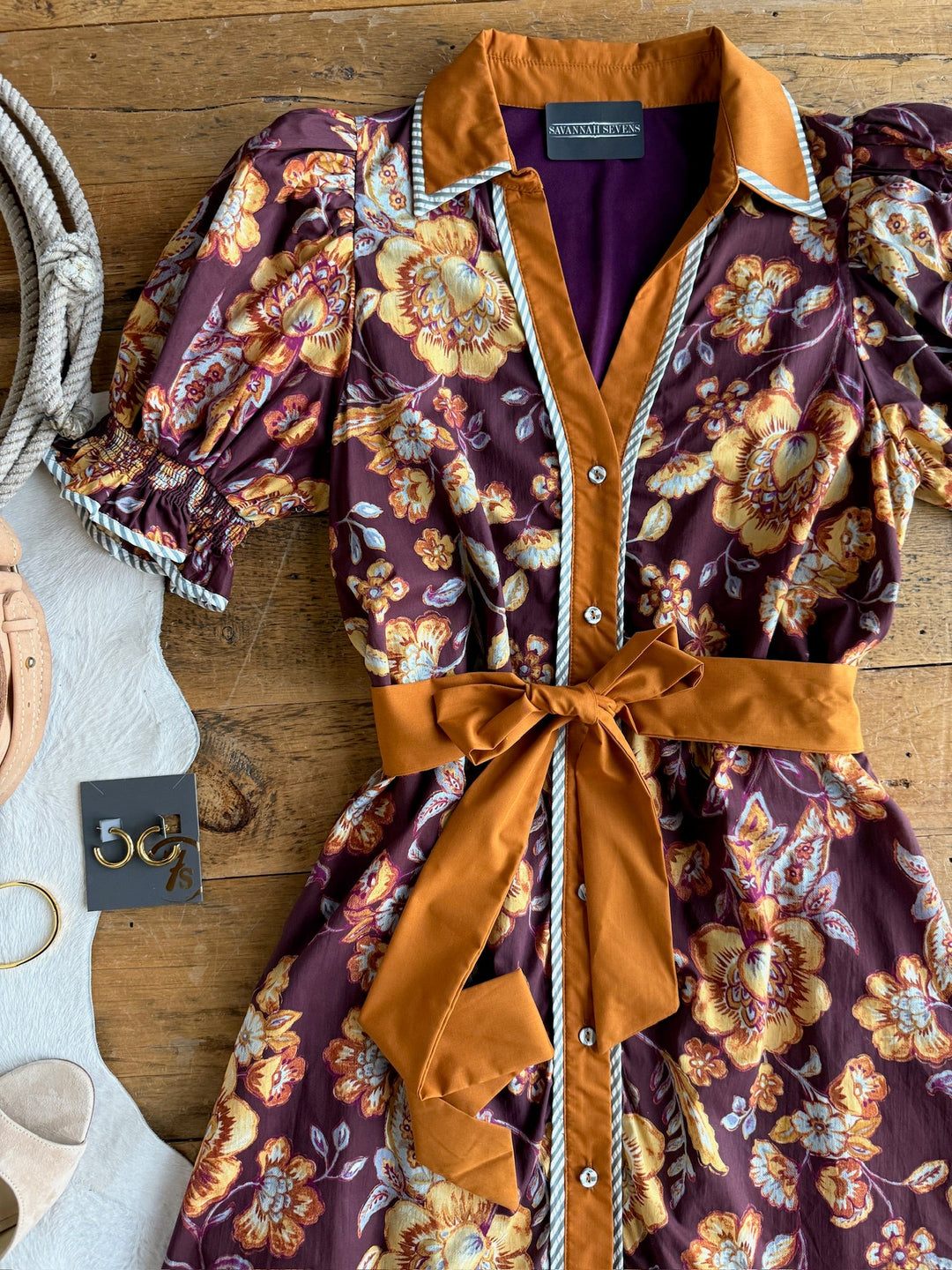 The Tullahassee Floral Dress by Savannah Sevens western life{&}style—a soft cotton midi dress with puffed sleeves, an orange collar, matching sash, and button-up front—is laid out on a wooden surface. It is displayed with sandals, a rope.