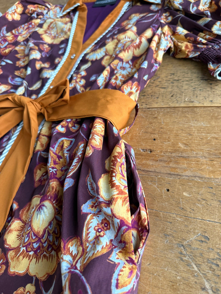 A close-up of the Tullahassee Floral Dress by Savannah Sevens western life{&}style, showcasing its yellow, orange, and purple floral patterns and a mustard-yellow sash, lying on a wooden surface.