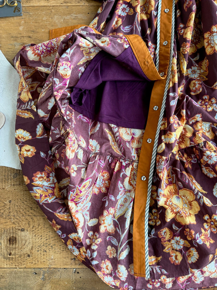 The Tullahassee Floral Dress by Savannah Sevens western life{&}style is displayed on a wooden surface, partially unbuttoned to showcase its purple inner lining. This button-up dress features delicate floral patterns in vibrant purple and orange hues, offering a fresh and stylish look.
