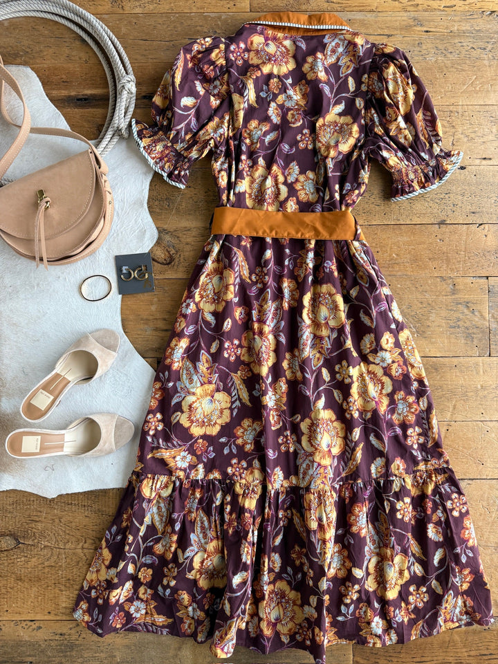The Tullahassee Floral Dress by Savannah Sevens western life{&}style, featuring puff sleeves and an orange belt, is styled with beige flats, a tan handbag, a bangle, and a pair of earrings, all arranged on a wooden floor and light rug.