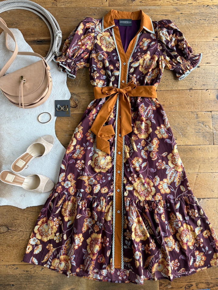The Tullahassee Floral Dress by Savannah Sevens western life{&}style—a button-up, puff sleeve midi with sash—is paired with cream flats, a tan handbag, round earrings, and sunglasses, all arranged on a wooden floor.