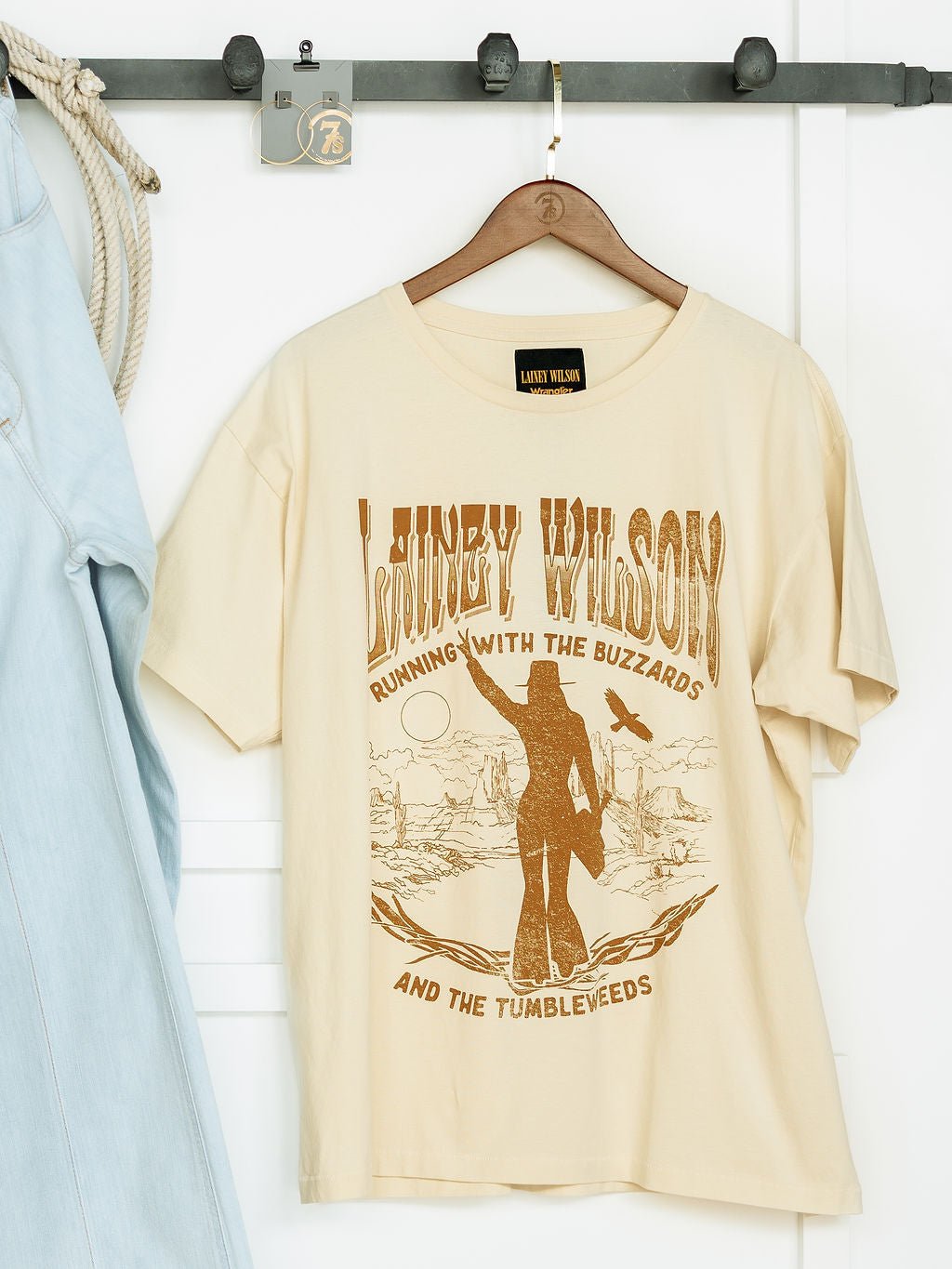 A beige Tumbleweed Graphic Tee by Savannah Sevens western life{&}style hangs on a hook next to a light denim Wrangler shirt, featuring Running With The Buzzards and The Tumbleweeds with a western-style woman and landscape graphic.