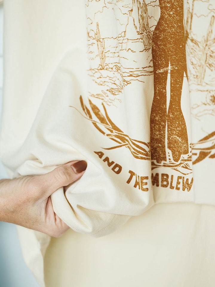 A hand with brown nail polish holds the Savannah Sevens western life{&}style "Tumbleweed Graphic Tee," a cream oversized cotton shirt featuring brown cactus illustrations and partially visible text AND THEMBLEW.