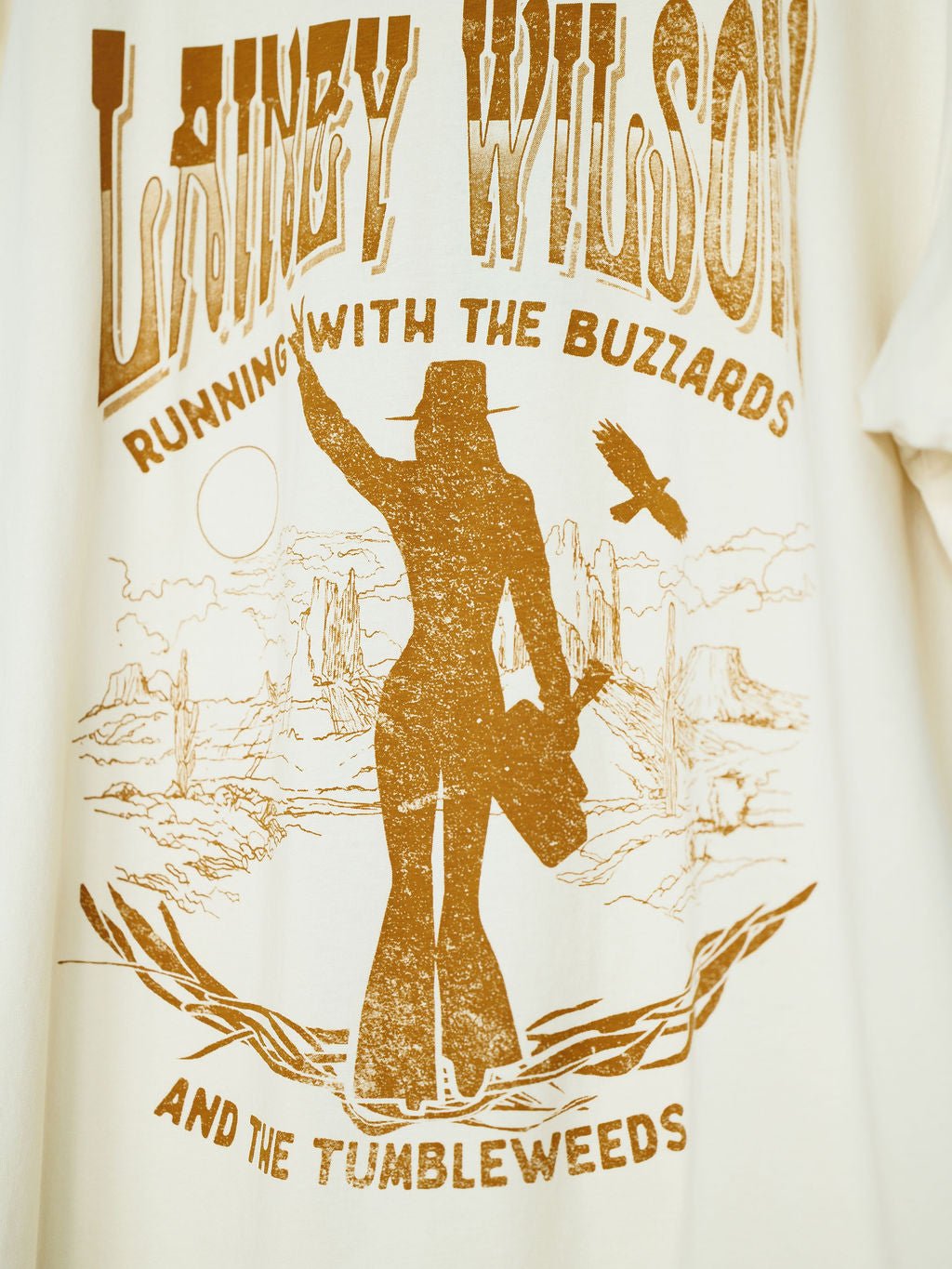 The Tumbleweed Graphic Tee by Savannah Sevens western life{&}style features an oversized cotton fit with a graphic of a woman in a cowboy hat, desert scene, buzzards, and text: Lainey Wilson—Running with the Buzzards and the Tumbleweeds.