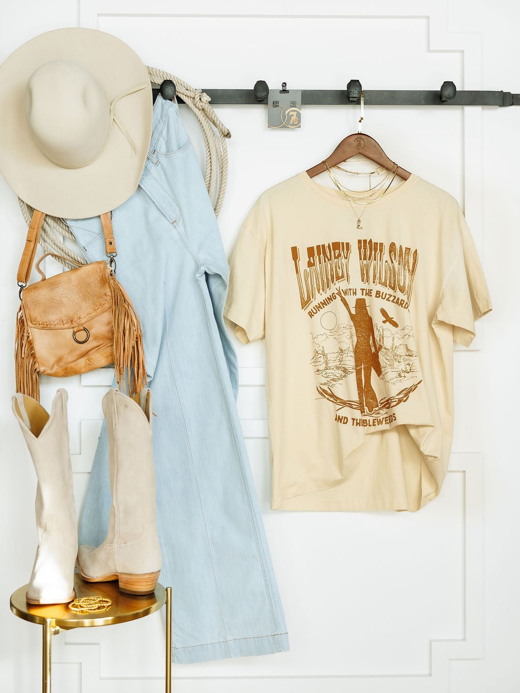 A beige cowboy hat, light blue denim overalls, a tan fringed purse, cream cowboy boots, and the Tumbleweed Graphic Tee by Savannah Sevens western life{&}style hang on hooks against a white wall, creating a Western-inspired look.
