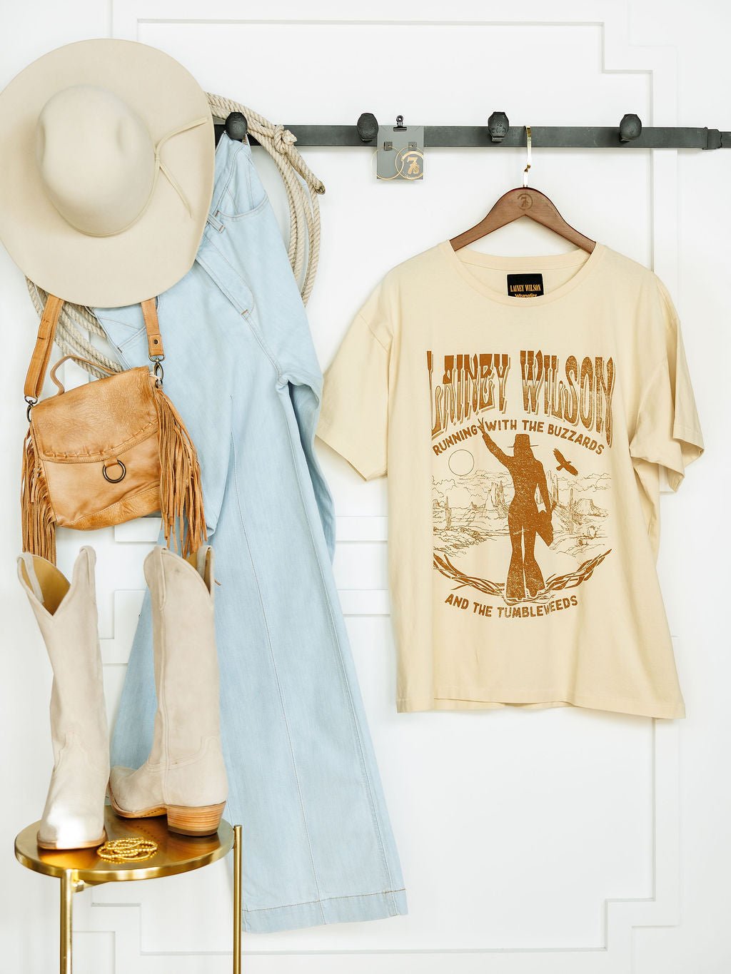 The cream Tumbleweed Graphic Tee by Savannah Sevens western life{&}style hangs on a hook beside a light blue denim jumpsuit, beige wide-brim hat, tan fringe purse, coiled rope, and beige cowboy boots on a small golden table.