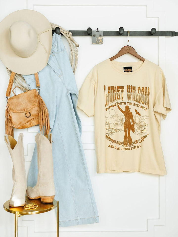The cream Tumbleweed Graphic Tee by Savannah Sevens western life{&}style hangs on a hook beside a light blue denim jumpsuit, beige wide-brim hat, tan fringe purse, coiled rope, and beige cowboy boots on a small golden table.