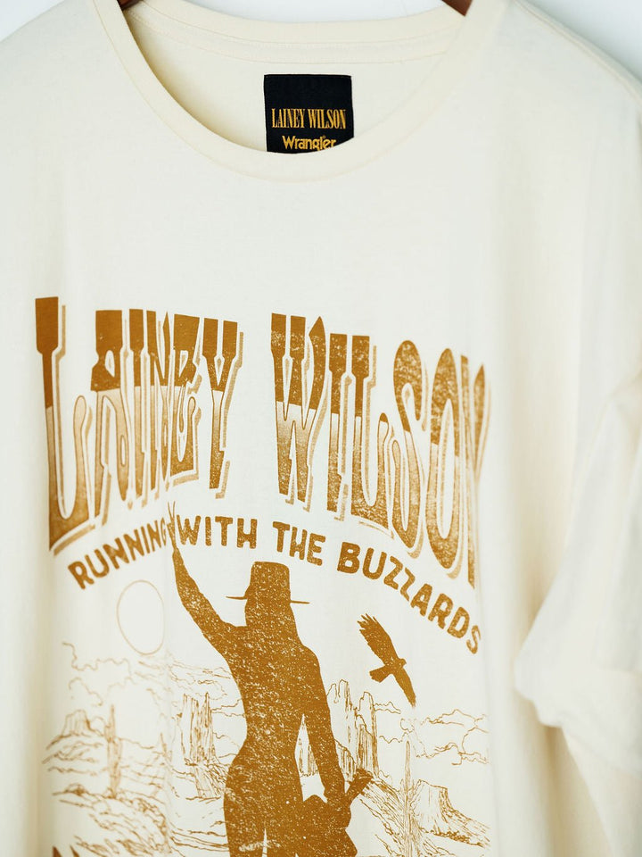 The Tumbleweed Graphic Tee by Savannah Sevens western life{&}style is a cream oversized cotton shirt with “LAINEY WILSON” and “RUNNING WITH THE BUZZARDS” in bold brown, plus cowgirl, buzzard, desert rock, and rising sun graphics.