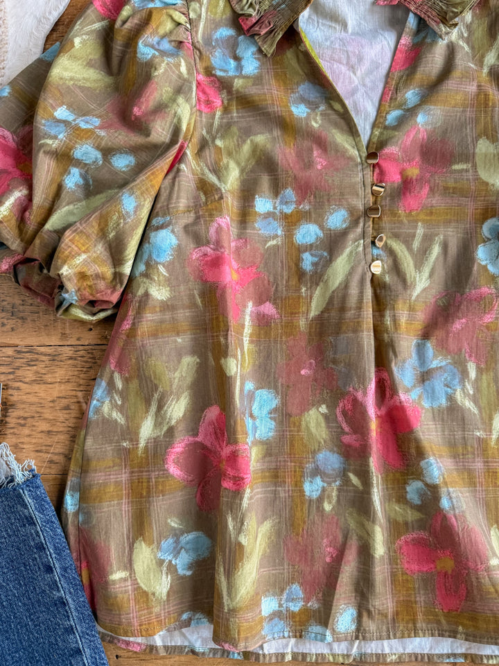 A close-up of the Tupelo Olive Puff Sleeve Top by Savannah Sevens western life{&}style, featuring cotton ruffles, puffy sleeves, gold buttons, and a soft pastel plaid pattern in pink, blue, and green, displayed next to a pair of blue jeans on a wooden surface.