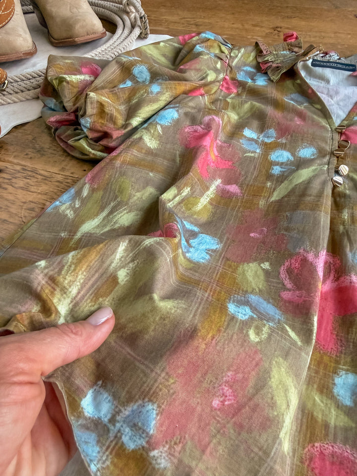 A hand holds the Tupelo Olive Puff Sleeve Top by Savannah Sevens western life{&}style—a sheer short sleeve top with a floral pattern in pink, blue, and green hues. In the background, boots, rope, and a white cloth rest on a wooden surface.