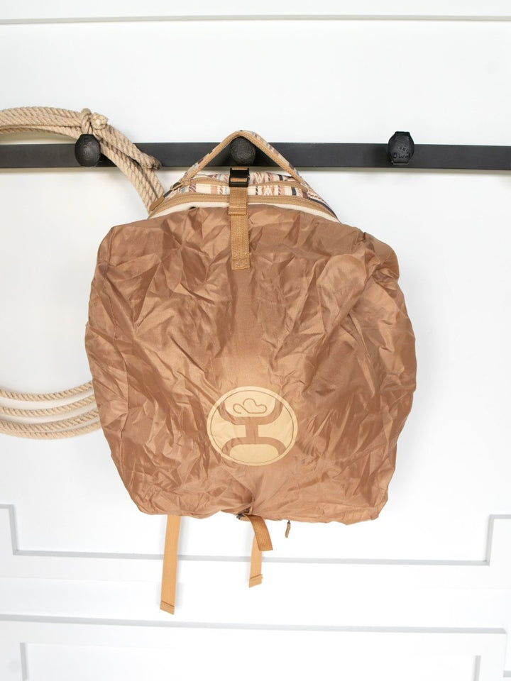 The Tuscola Medium Southwest Canvas Backpack by Savannah Sevens western life{&}style, featuring a brown rain cover and circular logo, hangs on a black wall hook beside a coiled beige rope against a white background.
