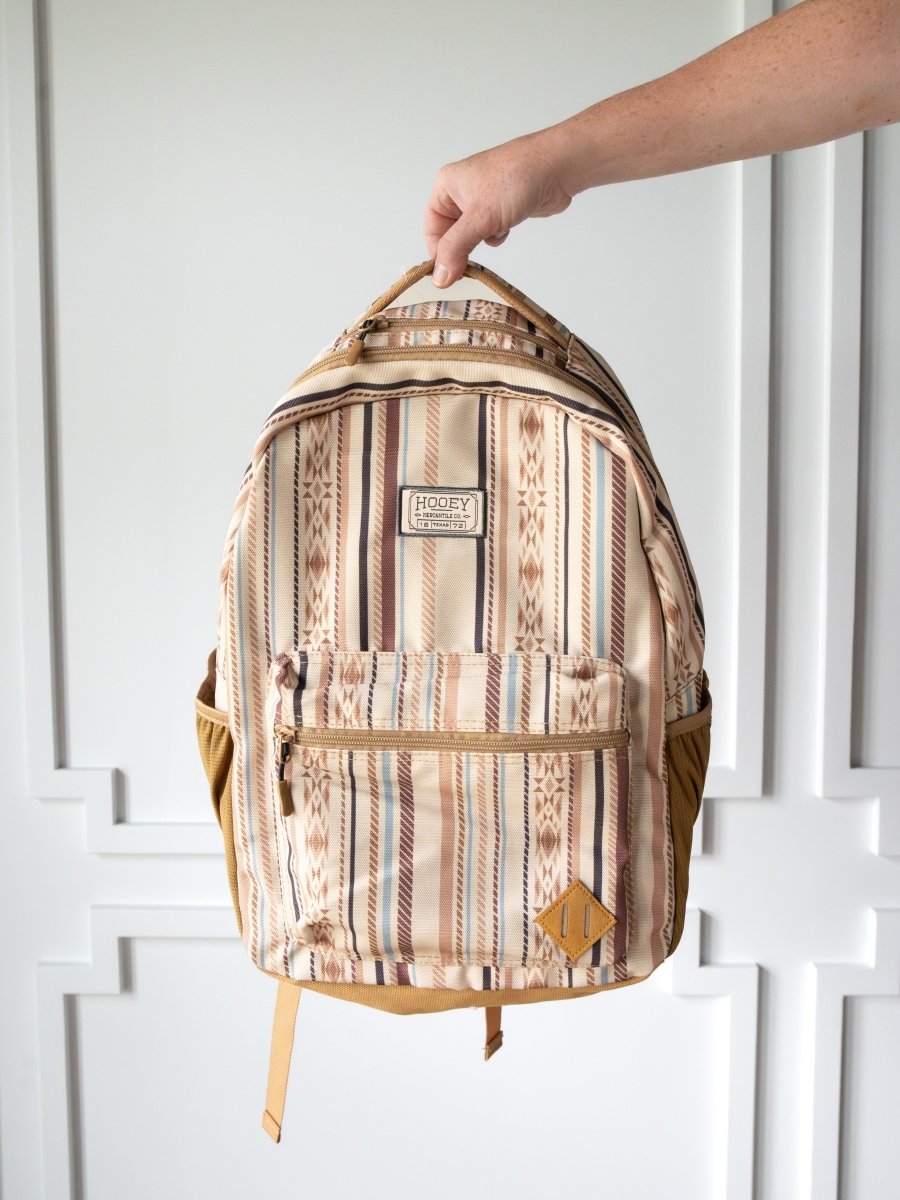 A hand holds the Tuscola Medium Southwest Canvas Backpack by Savannah Sevens western life{&}style, featuring brown, tan, and maroon southwest stripes, padded laptop sleeve, front zipper pocket, side pockets, and a logo patch.