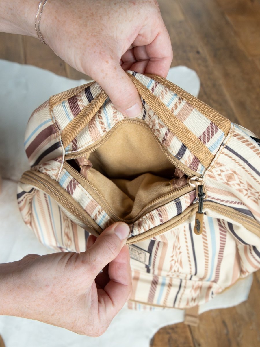 Someone holds open the Tuscola Medium Southwest Canvas Backpack by Savannah Sevens western life{&}style, revealing its empty interior, zippered pocket, and padded laptop sleeve against a wooden floor background.