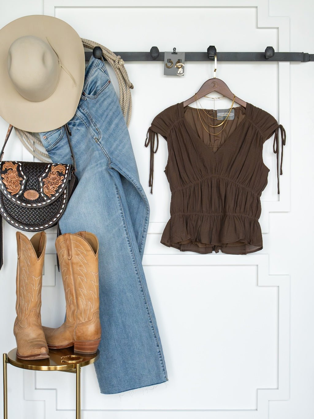 The Tustin Chocolate Ruched Top by Savannah Sevens western life{&}style, paired with a cowboy hat, jeans, a tooled leather purse, and tan cowboy boots, are neatly arranged against a white wall—evoking a Western-inspired fashion style with young contemporary sizing.