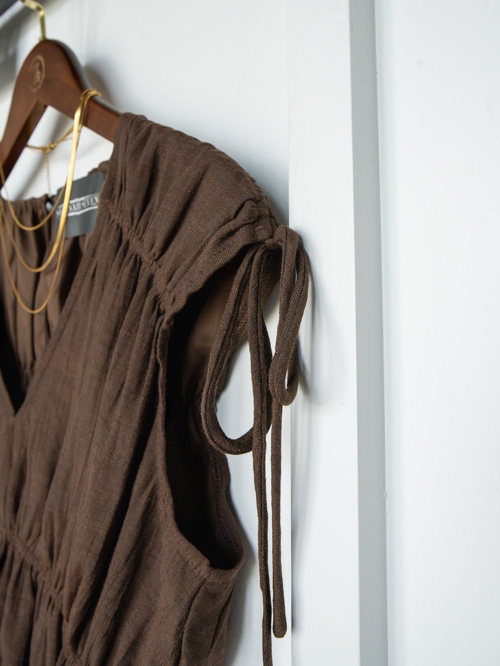 Close-up of the Tustin Chocolate Ruched Top by Savannah Sevens western life{&}style, featuring ruched elastic seams and gathered shoulder ties, displayed on a wooden hanger next to a gold necklace against a white wall.