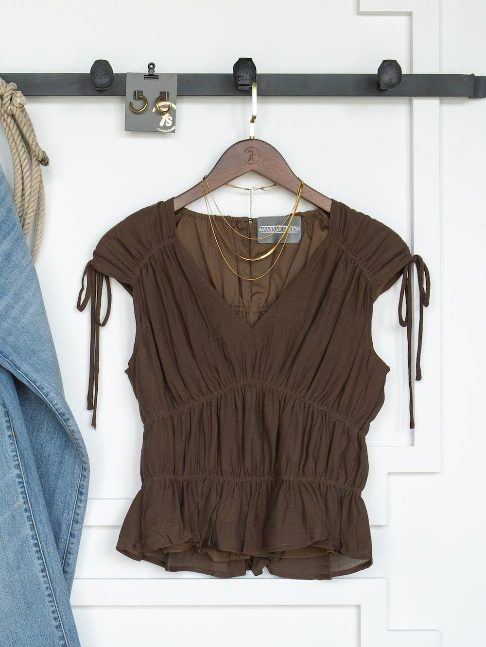 The Tustin Chocolate Ruched Top by Savannah Sevens western life{&}style, featuring cap sleeves and ruched elastic seams, hangs on a wooden hanger. Gold layered necklaces are draped over the hanger, while a pair of blue jeans is visible on the left.