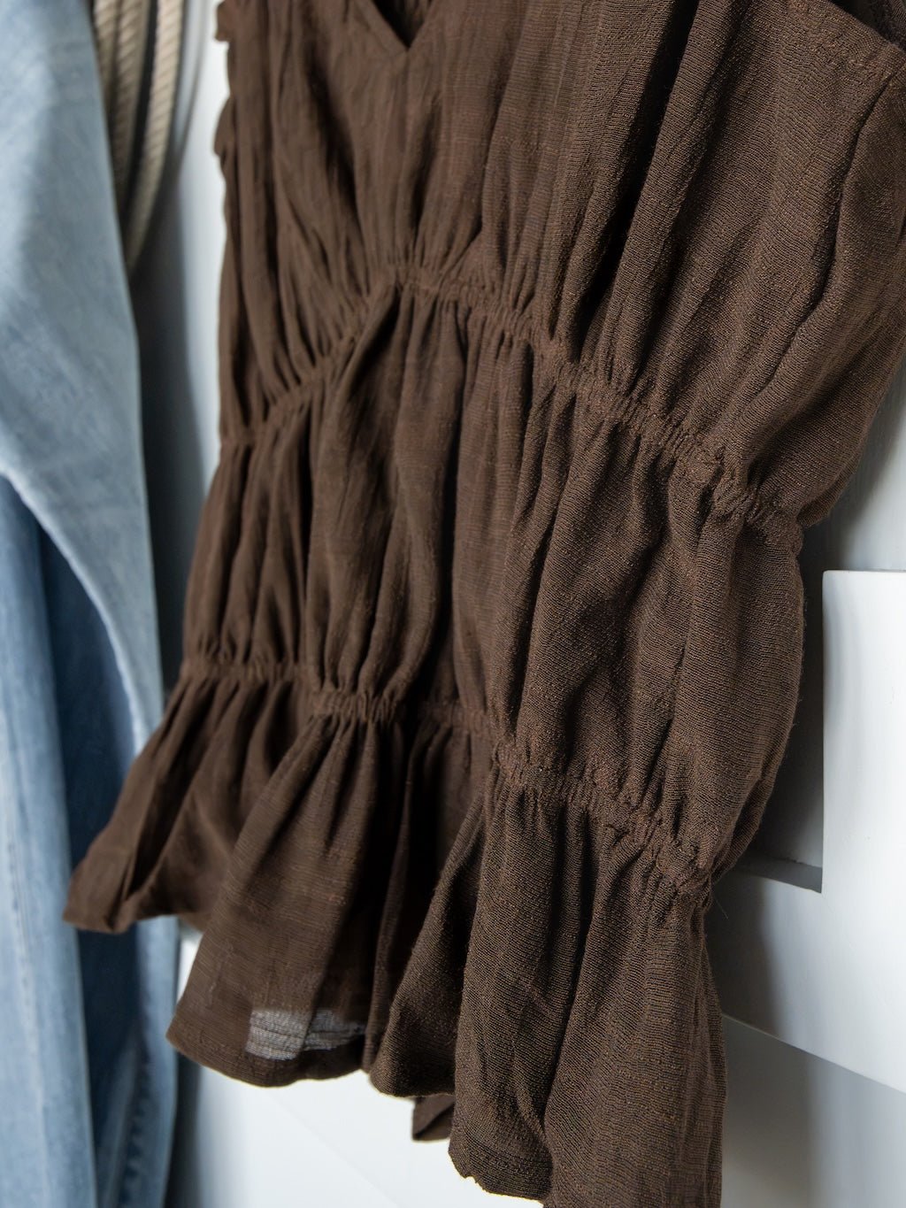 A close-up of the Savannah Sevens western life{&}style Tustin Chocolate Ruched Top—a brown, long-sleeve blouse featuring ruched detailing, ruffled cuffs, and elastic seams—hanging next to a light blue garment and highlighting young contemporary sizing.