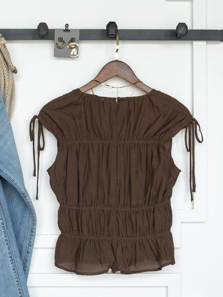 The Tustin Chocolate Ruched Top by Savannah Sevens western life{&}style, featuring cap sleeves, ruched elastic seams, and a ruffled hem, hangs on a wooden hanger next to a denim jacket against a white wall. Designed in young contemporary sizing.