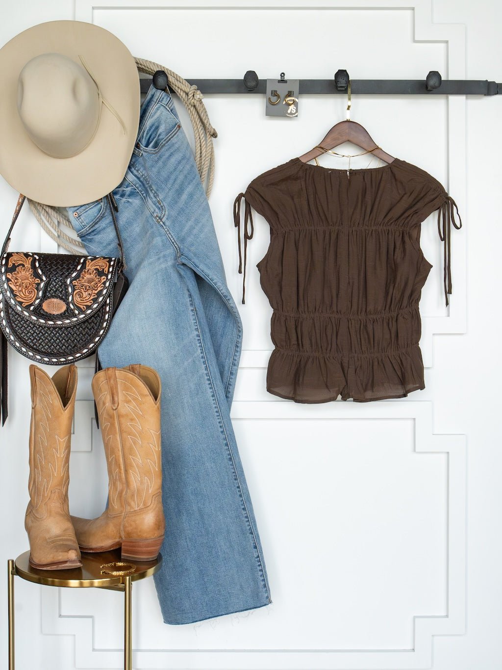 A beige hat, blue jeans, the Savannah Sevens western life{&}style Tustin Chocolate Ruched Top, a western-style purse, and cowboy boots are displayed neatly against a white wall in young contemporary sizing.