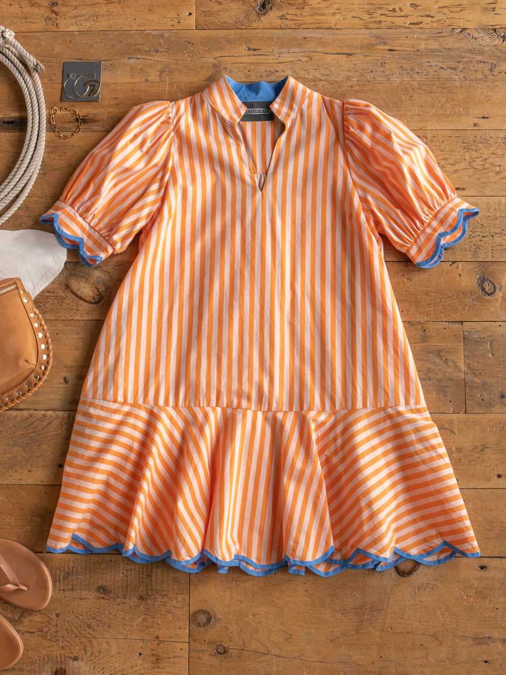 The Tyndall Orange Striped Puff Sleeve Dress by Savannah Sevens western life{&}style, featuring blue trim on the sleeves, collar, and hem, is displayed flat on a wooden surface with chic accessories like sandals and a handbag.