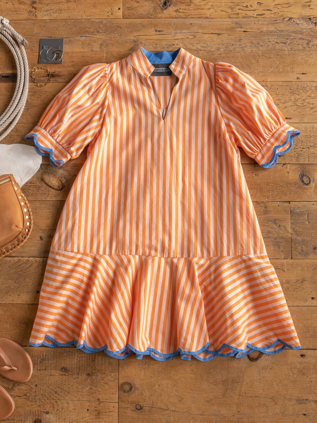 The Tyndall Orange Striped Puff Sleeve Dress by Savannah Sevens western life{&}style, featuring blue trim on the sleeves, collar, and hem, is displayed flat on a wooden surface with chic accessories like sandals and a handbag.