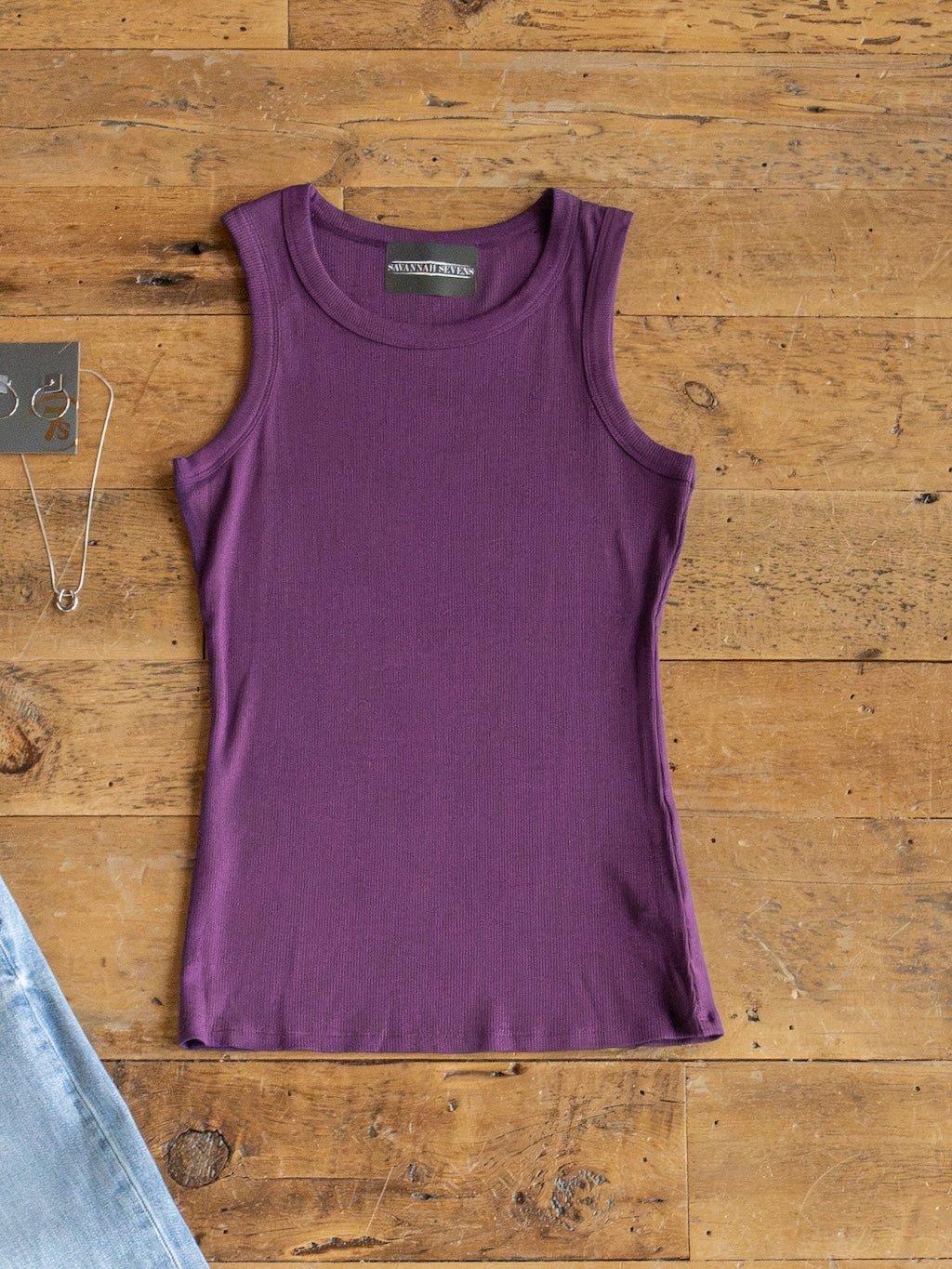 The Utica Plum Ribbed Basic Tank by Savannah Sevens western life{&}style is displayed laid flat on a wooden surface, paired with a silver necklace and partially visible light blue jeans in the corners.