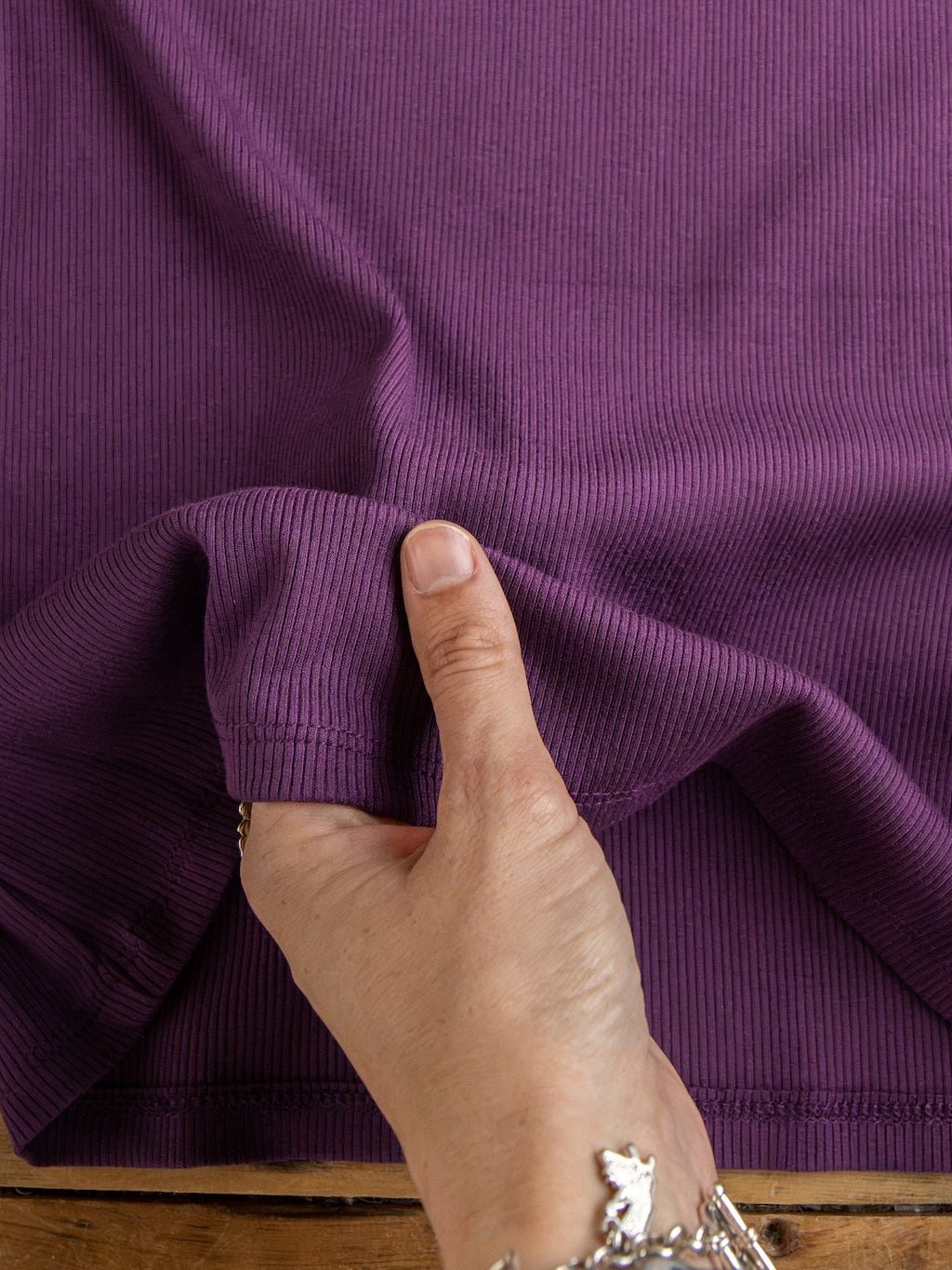 A hand lightly pinches and lifts a section of ribbed purple fabric from the Utica Plum Ribbed Basic Tank by Savannah Sevens western life{&}style, highlighting the texture and stretch of this women's contemporary top. The person wears a silver charm bracelet, with a wooden surface in the background.