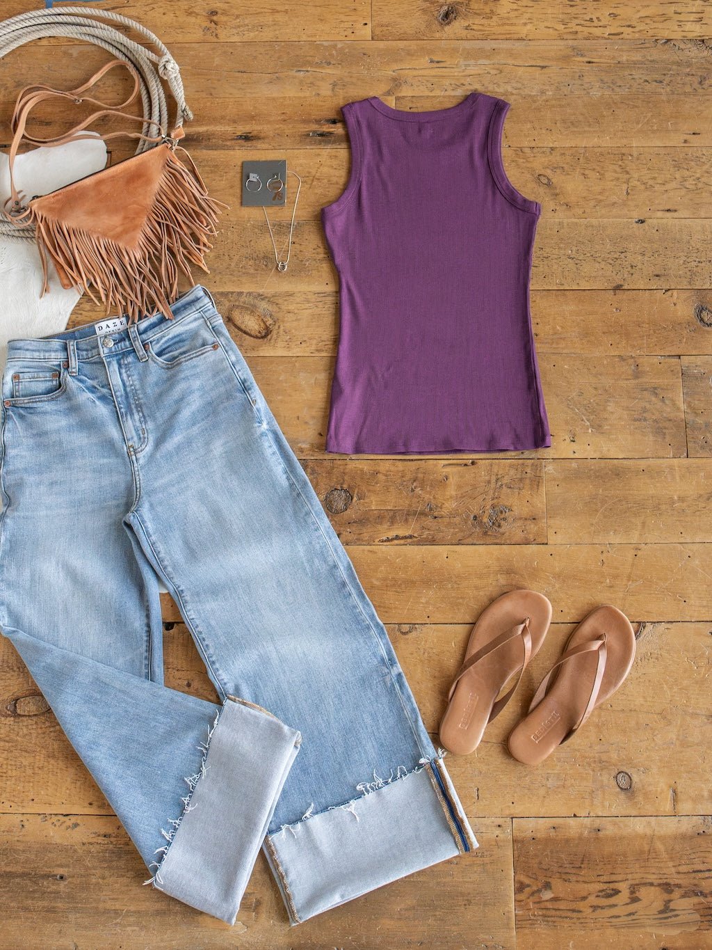 The Utica Plum Ribbed Basic Tank by Savannah Sevens western life{&}style, paired with light-wash jeans, tan flip-flops, a tan fringe purse, a white bag, a necklace, and earrings are displayed on a wooden floor.