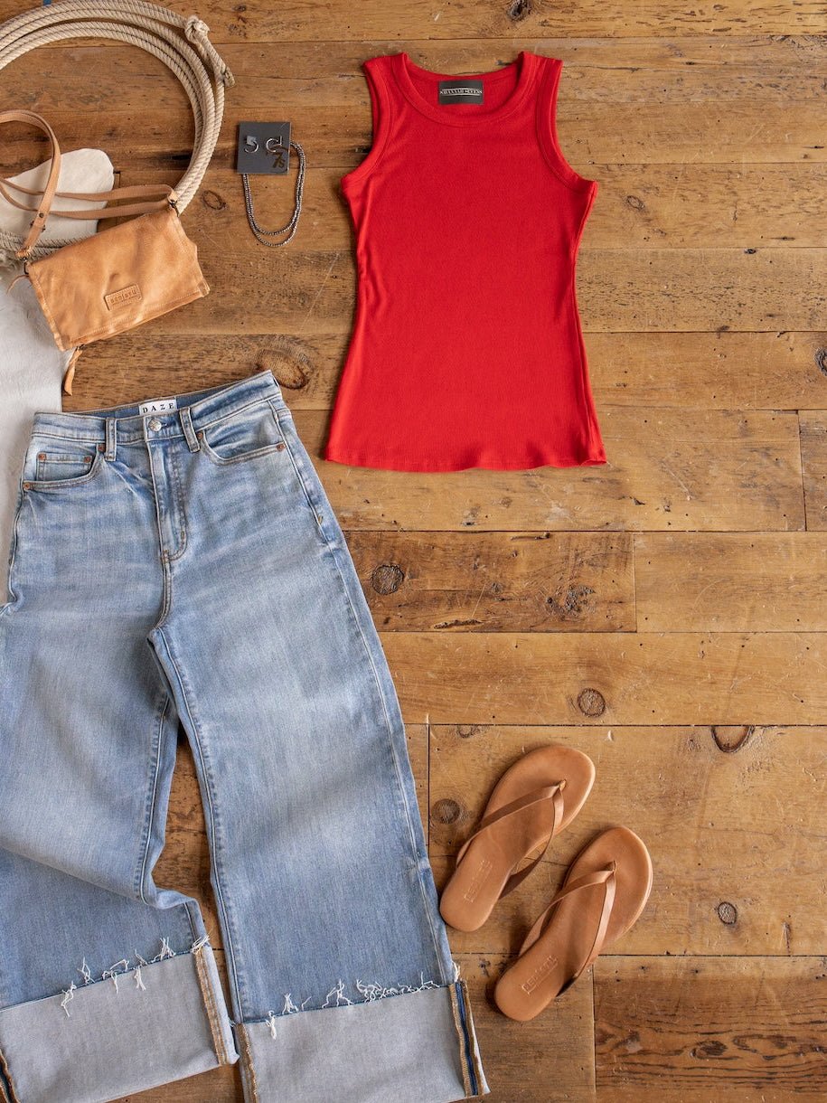 Flat lay featuring the Utica Red Ribbed Basic Tank from Savannah Sevens western life{&}style, paired with light blue cuffed jeans, tan flip-flops, a tan crossbody bag, sunglasses, and a necklace on a wooden floor.