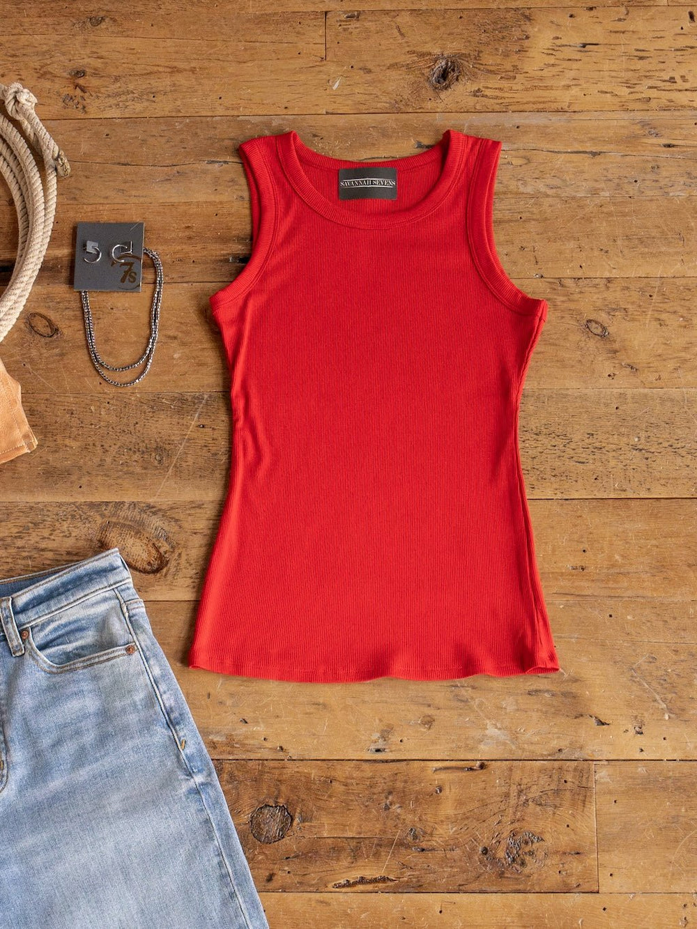 The Utica Red Ribbed Basic Tank by Savannah Sevens western life{&}style is laid flat on a rustic wooden surface beside light blue jeans, a silver necklace, and a metal accessory.