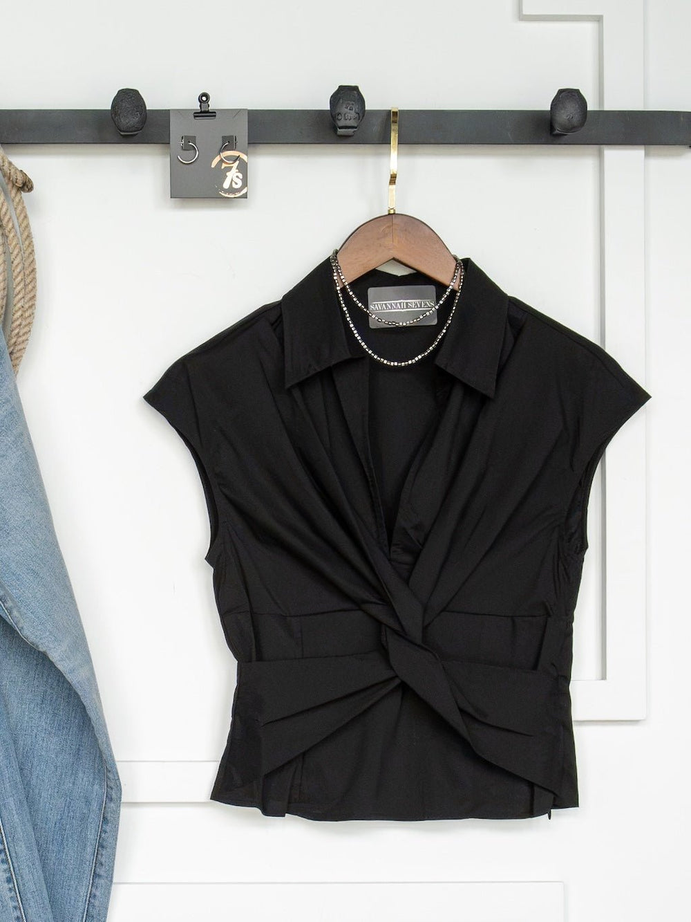 The Valencia Black Twist Front Top by Savannah Sevens western life{&}style hangs on a wooden hanger against a white wall with hooks. A necklace is draped over the collar, and blue denim is partially visible on the left side.