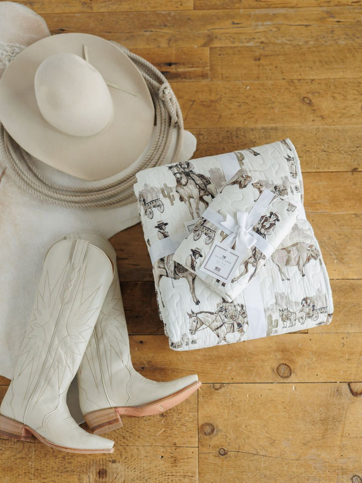 A cream cowboy hat, coiled rope, white boots, and the Vaquera Quilt & Shams by Savannah Sevens western life&style—with a matching gift-wrapped set—rest on a wooden floor.