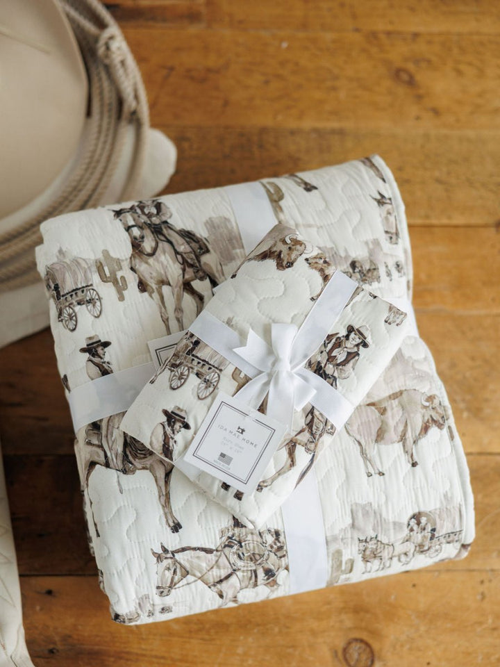 The Vaquera Quilt & Shams by Savannah Sevens western life{&}style, featuring a cowboy, horse, and wagon western print, are shown with a coordinating gift package tied with a white ribbon and tag on a wooden surface with coiled rope nearby.