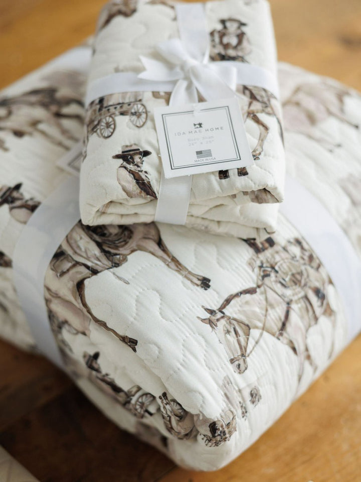 The Vaquera Quilt & Shams by Savannah Sevens western life{&}style, featuring a cowboy and horse print, is displayed folded with a white ribbon on wood. This handmade 100% cotton quilt offers comfort and western style.