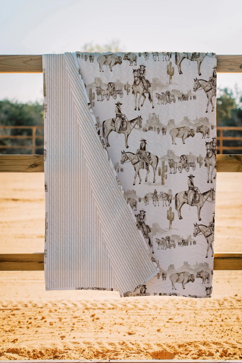 The Vaquera Quilt & Shams by Savannah Sevens western life{&}style hangs on a wooden fence, showcasing a cowboy, horse, and cactus print on one side and thin stripes on the other, all in 100% cotton against a sandy outdoor backdrop.