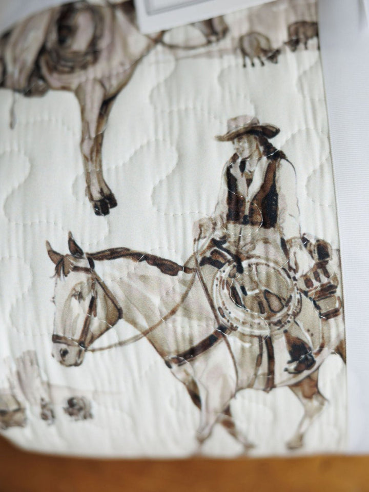 The Vaquera Quilt & Shams by Savannah Sevens western life{&}style features 100% cotton fabric with a western print of a cowboy in hat and vest on a saddled horse in brown and beige tones atop a white background.