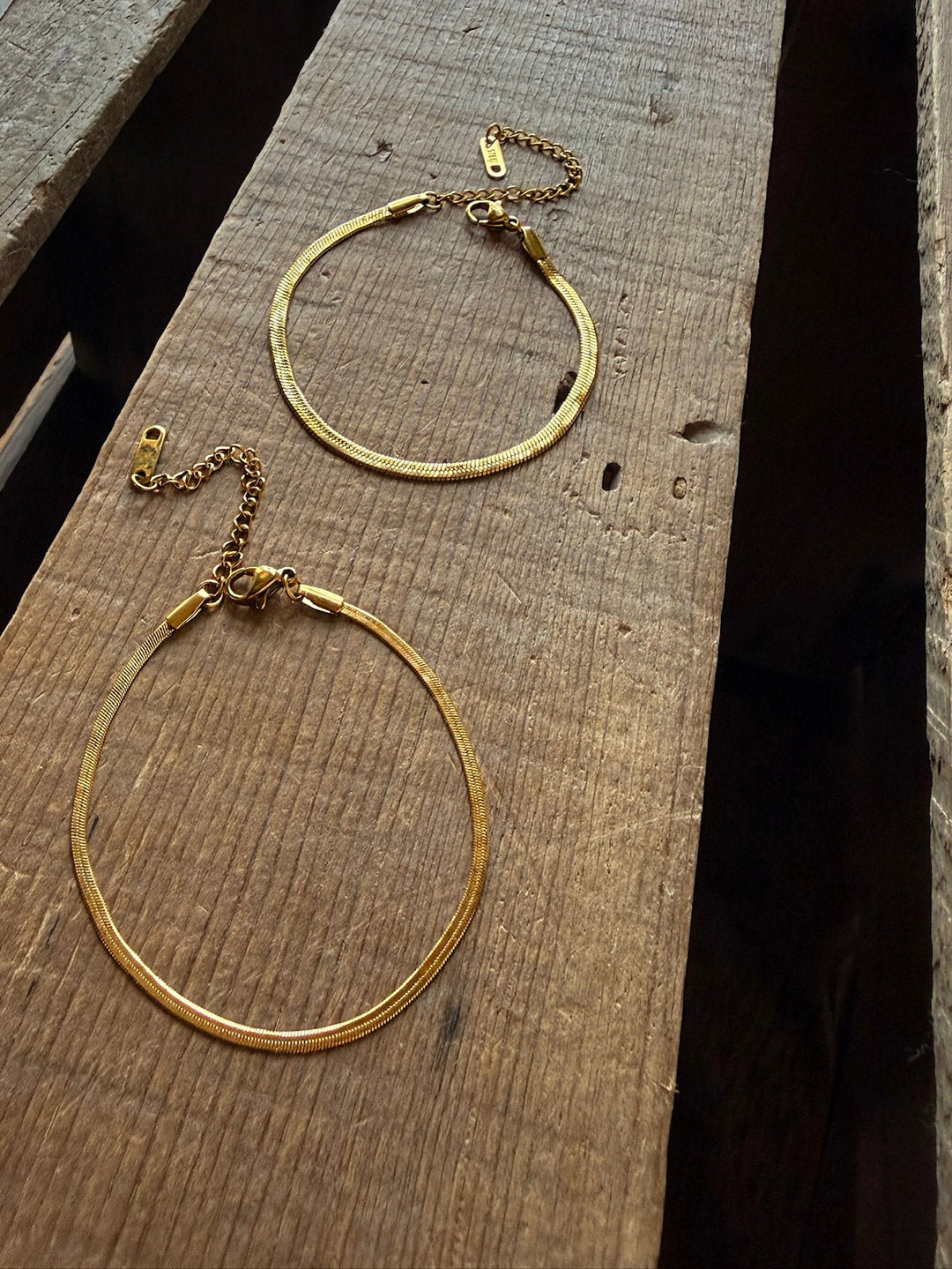 Two gold chain necklaces with clasps are laid out parallel on a rustic wooden surface, their texture and the gaps between the wood planks clearly visible. Beside them rests the Vera Gold Snake Bracelet by Savannah Sevens western life{&}style—a sleek, waterproof, and hypoallergenic accessory.