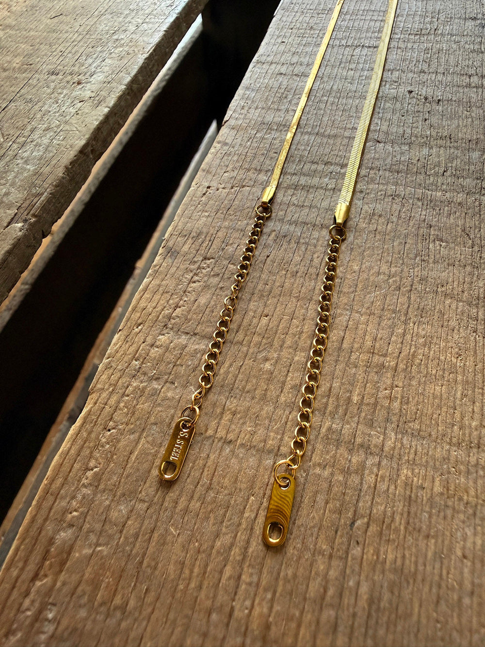 Two gold chain necklaces with rectangular tags at the ends rest side by side on a wooden surface in natural light, beautifully complementing the elegance of the Vera Gold Snake Bracelet by Savannah Sevens western life{&}style.