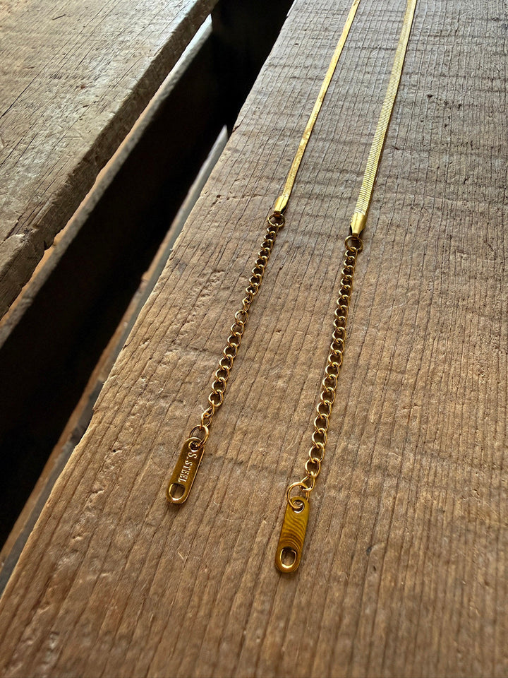 Two gold chain necklaces with rectangular tags at the ends rest side by side on a wooden surface in natural light, beautifully complementing the elegance of the Vera Gold Snake Bracelet by Savannah Sevens western life{&}style.