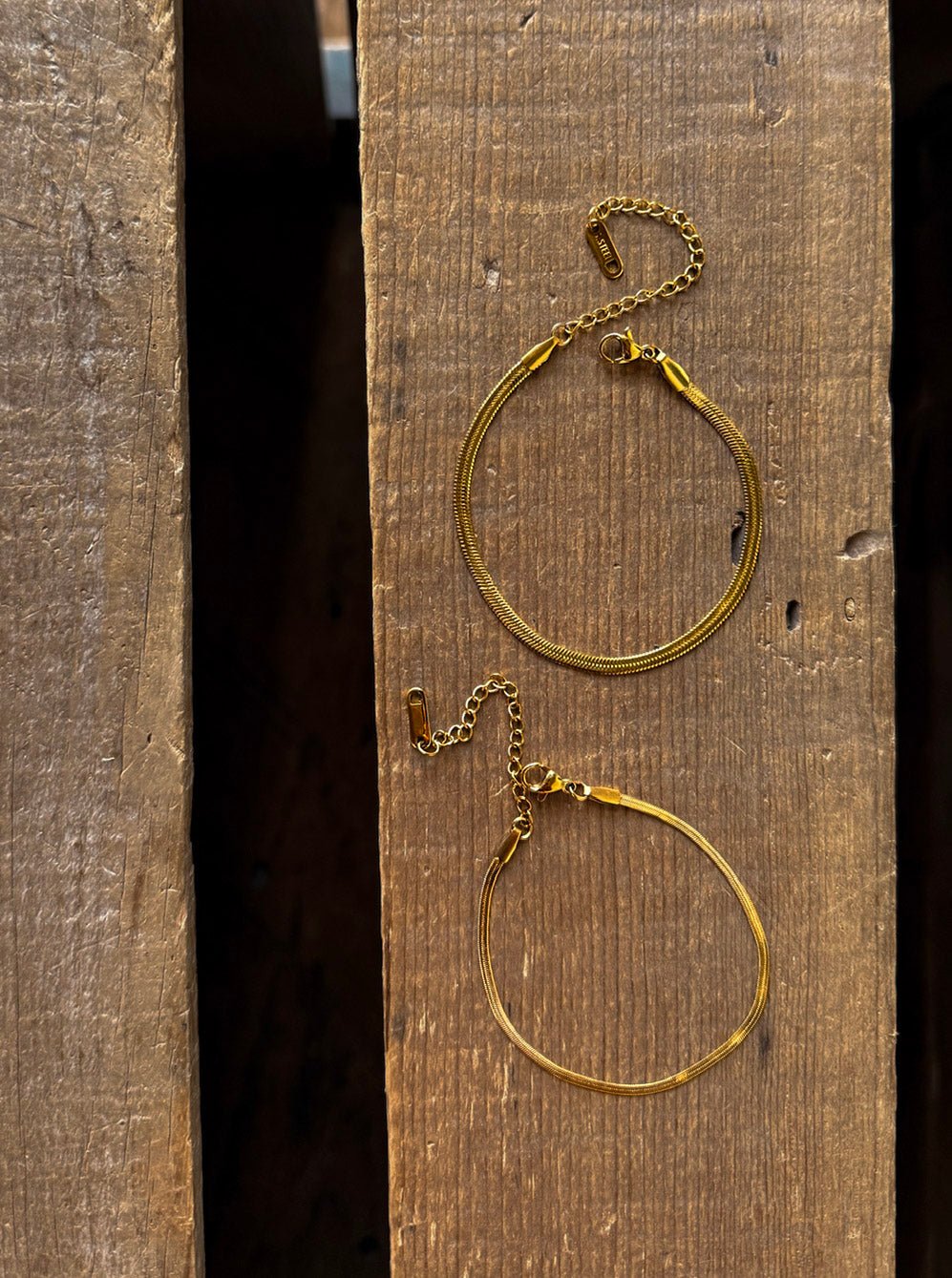 Two delicate gold chain bracelets, including the Vera Gold Snake Bracelet by Savannah Sevens western life{&}style featuring adjustable clasps, are positioned one above the other on a rustic wooden surface, displayed parallel to the wood grain. These hypoallergenic pieces from Savannah Sevens offer both style and comfort.