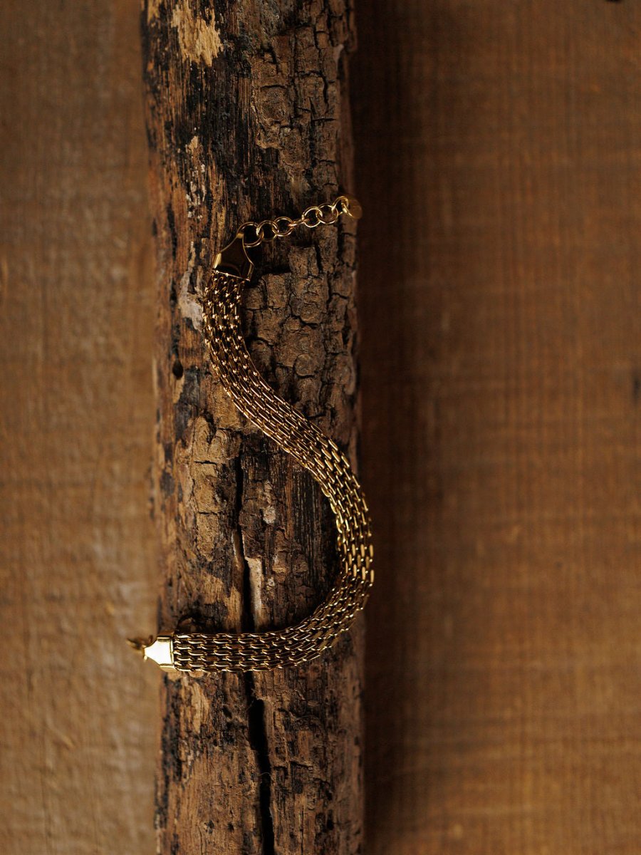 The Vienna Bracelet by Savannah Sevens western life{&}style features a gold braided S-shape design, resting on a textured wooden surface with a warm brown background. This 18K gold-filled piece is hypoallergenic.