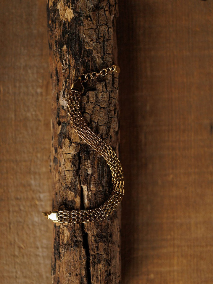 The Vienna Bracelet by Savannah Sevens western life{&}style features a gold braided S-shape design, resting on a textured wooden surface with a warm brown background. This 18K gold-filled piece is hypoallergenic.
