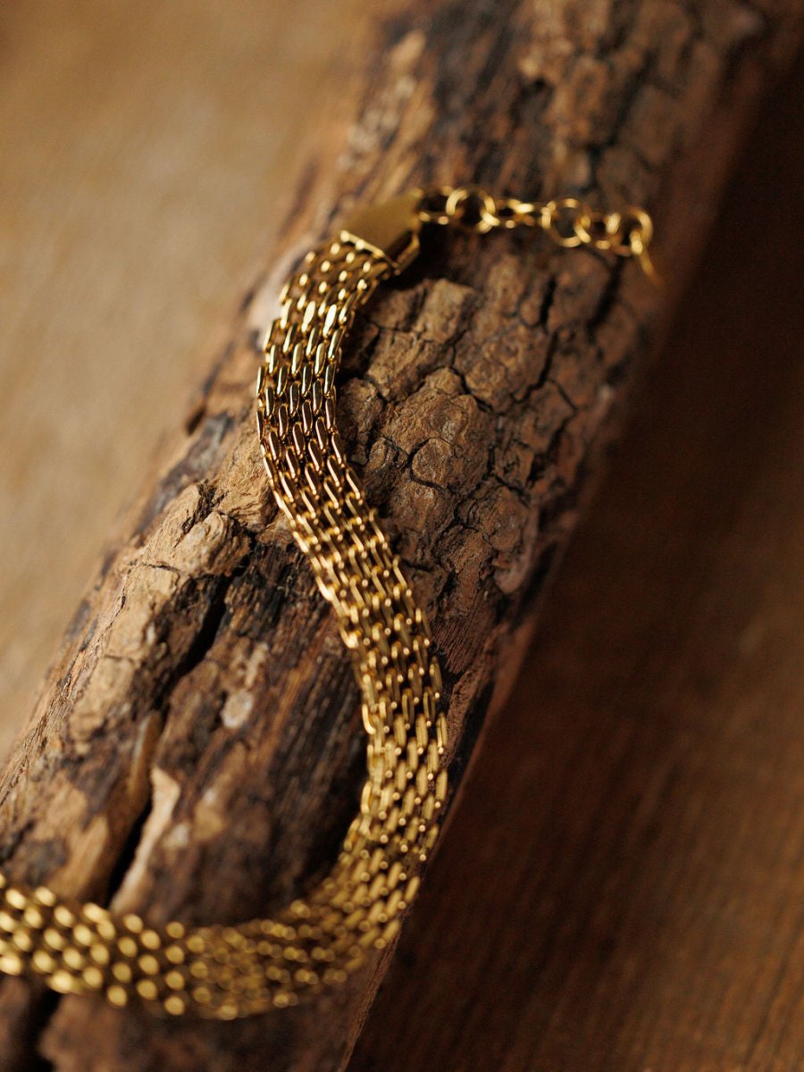 The Vienna Bracelet by Savannah Sevens western life{&}style—a hypoallergenic, woven gold braided design—rests on textured wood, showcasing its intricate links and polished 18K gold-filled finish.